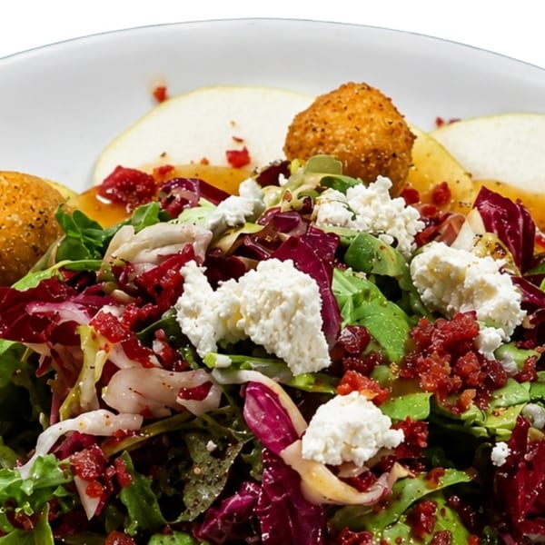 A fresh salad with mixed greens, crumbled cheese, bacon bits, fried balls, and pear slices.