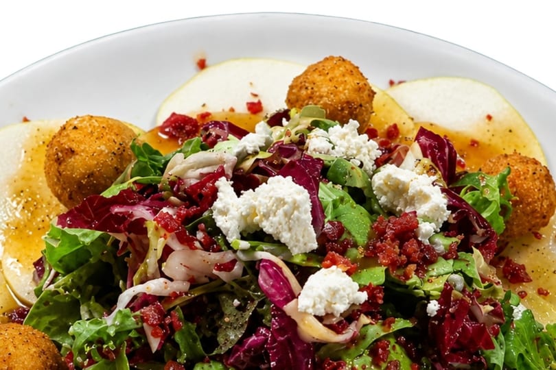 A fresh salad with mixed greens, crumbled cheese, bacon bits, fried balls, and pear slices.
