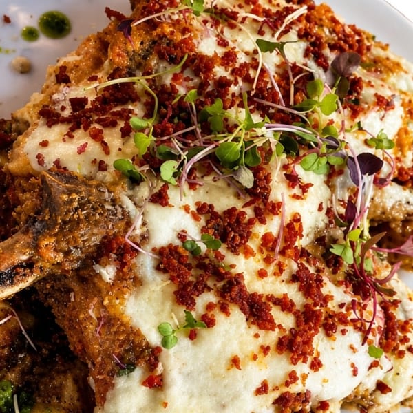 Two bone-in pork chops covered in melted cheese, bacon bits, and microgreens, with green sauce.