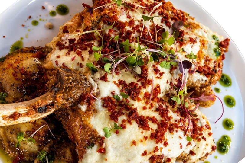 Two bone-in pork chops covered in melted cheese, bacon bits, and microgreens, with green sauce.