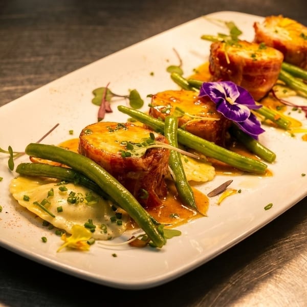 Plated dish with bacon-wrapped scallops, green beans, ravioli, and a purple edible flower.