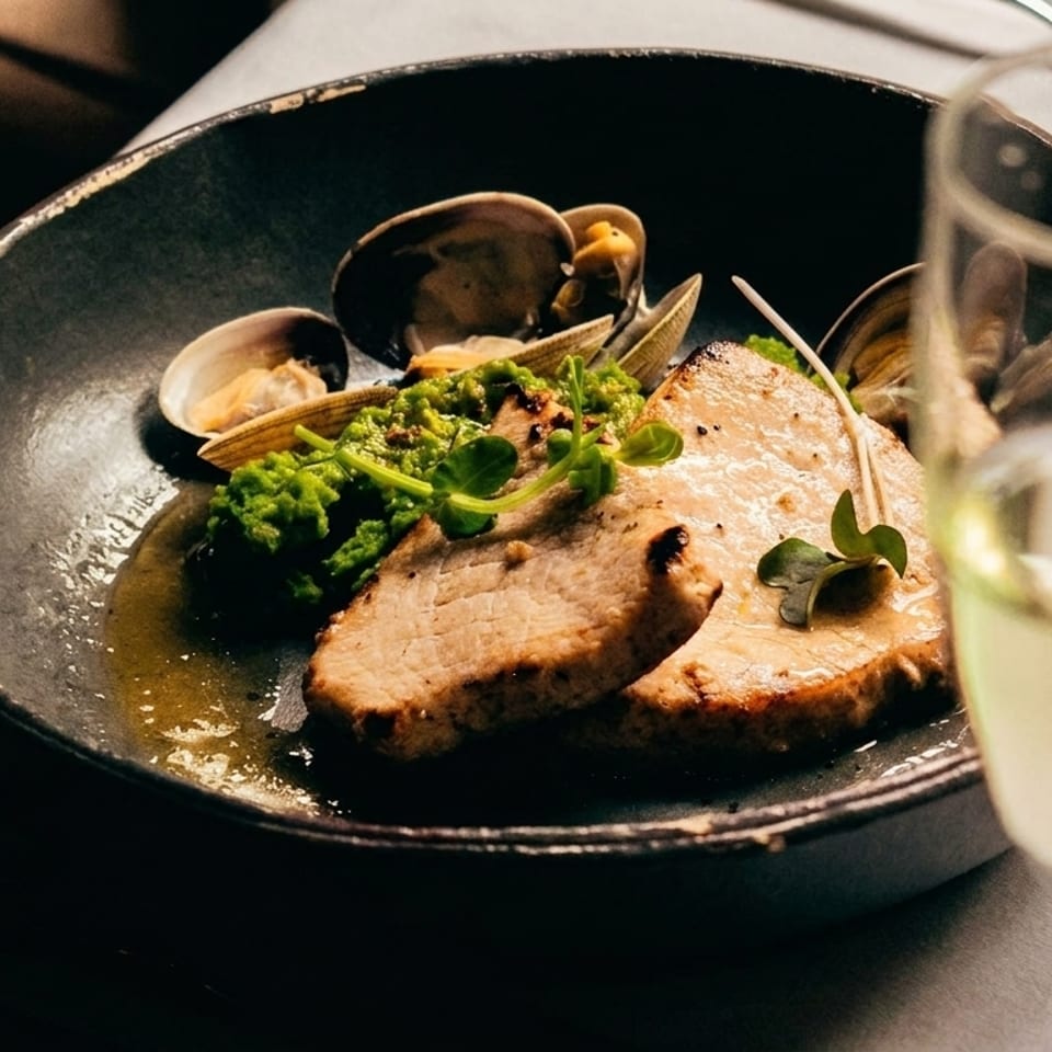 Seared pork slices with clams, green vegetables, and a glass of white wine on a table.