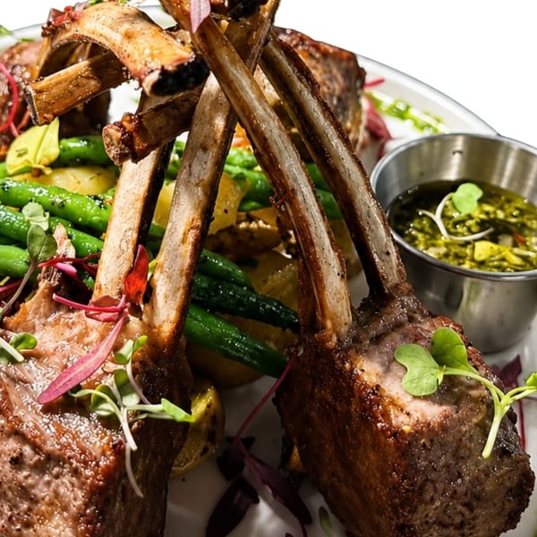 Grilled lamb chops with green beans, potatoes, and a side of pesto sauce on a white plate.